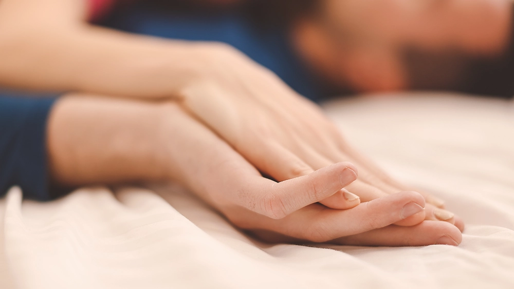 Main de femme caressant celle de son partenaire sur un lit, illustrant la découverte du corps de l’autre et des zones érogènes.