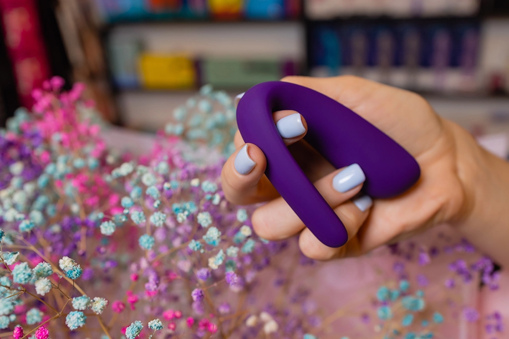 Main présentant un sextoy de couple dans un love store avec fleurs séchées et rayons en fond.